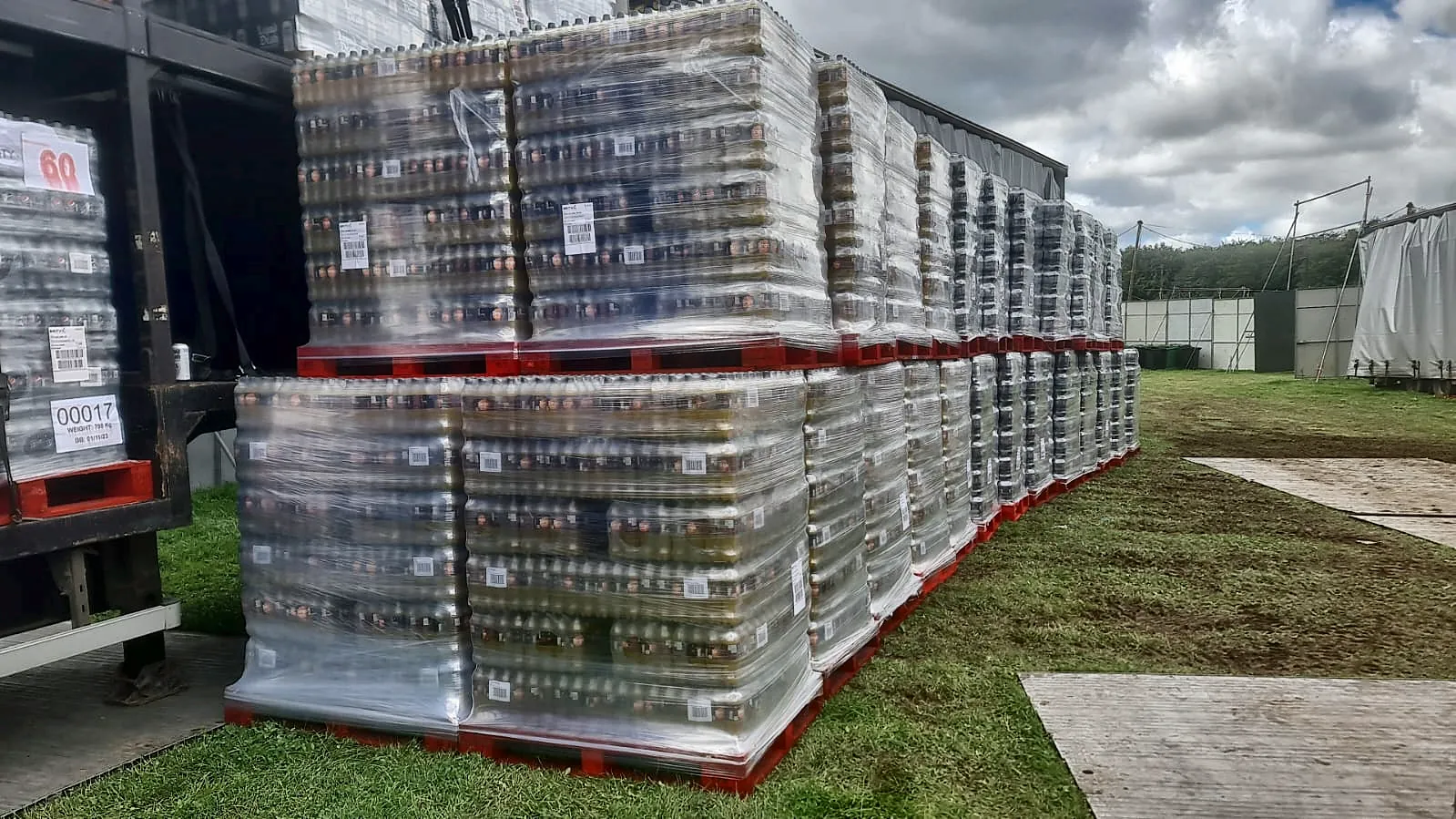 Double-stacked pallets of soft drinks lined up beside a trailer on grass